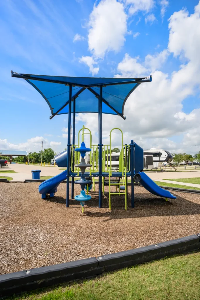 Blue and lime-green children's play structure with two slides under a blue shade canopy on wood mulch, RVs parked in